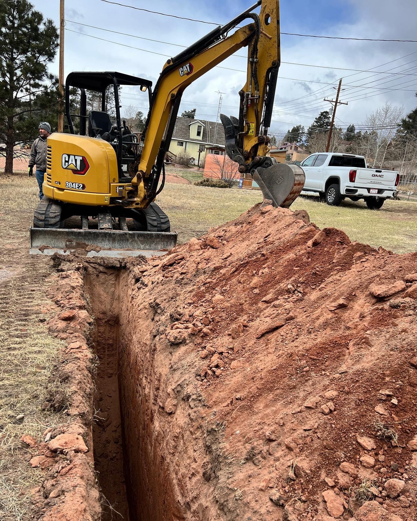 Excavator digging a trench in a yard with trees and vehicles in the background.