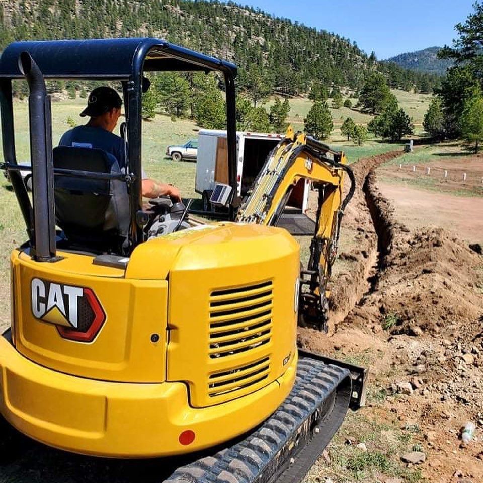 Person operating a yellow excavator digging a trench in a grassy landscape with trees and hills.