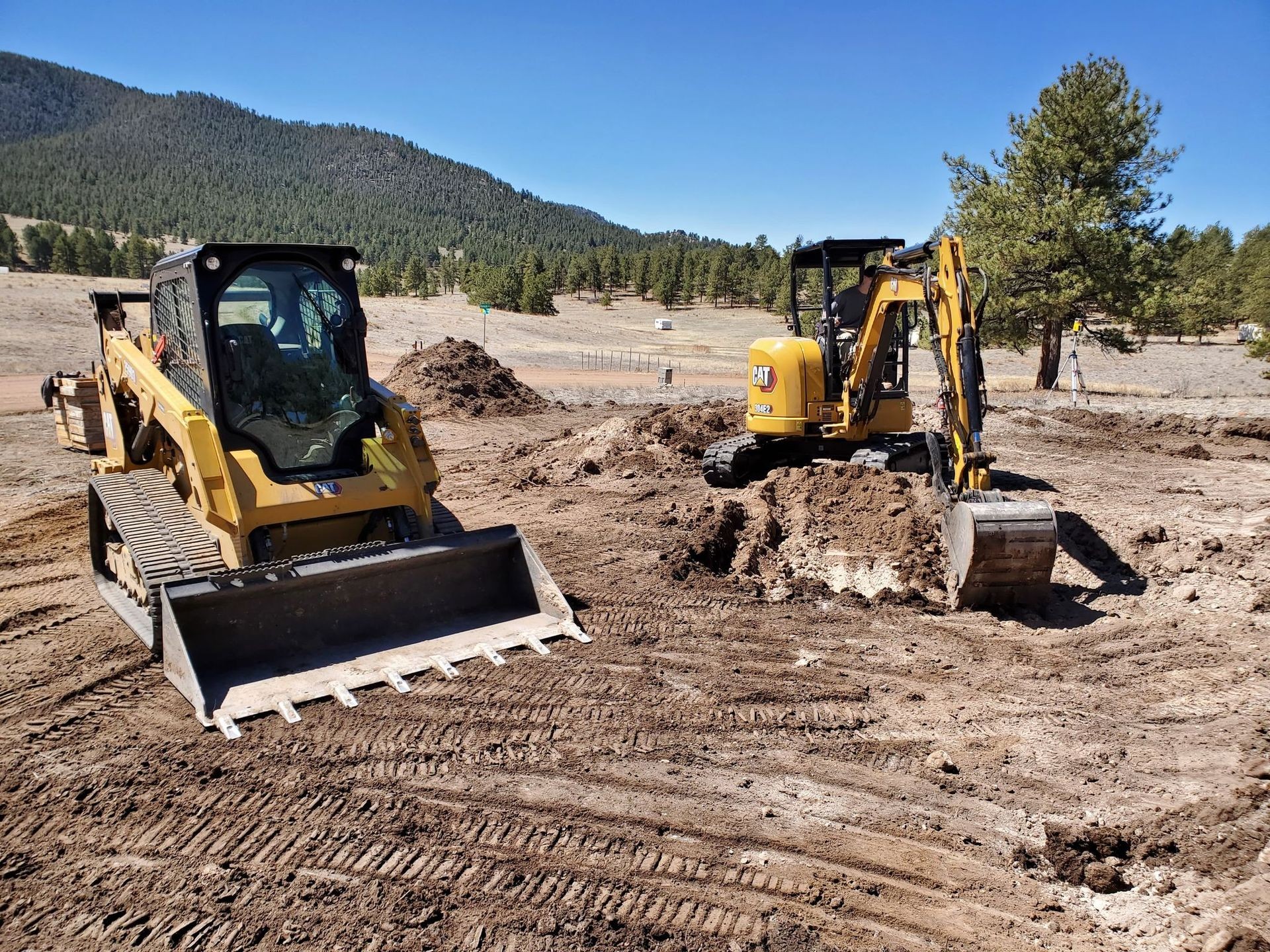 Two yellow construction vehicles digging dirt on a sunny day, with mountains and trees in the background.